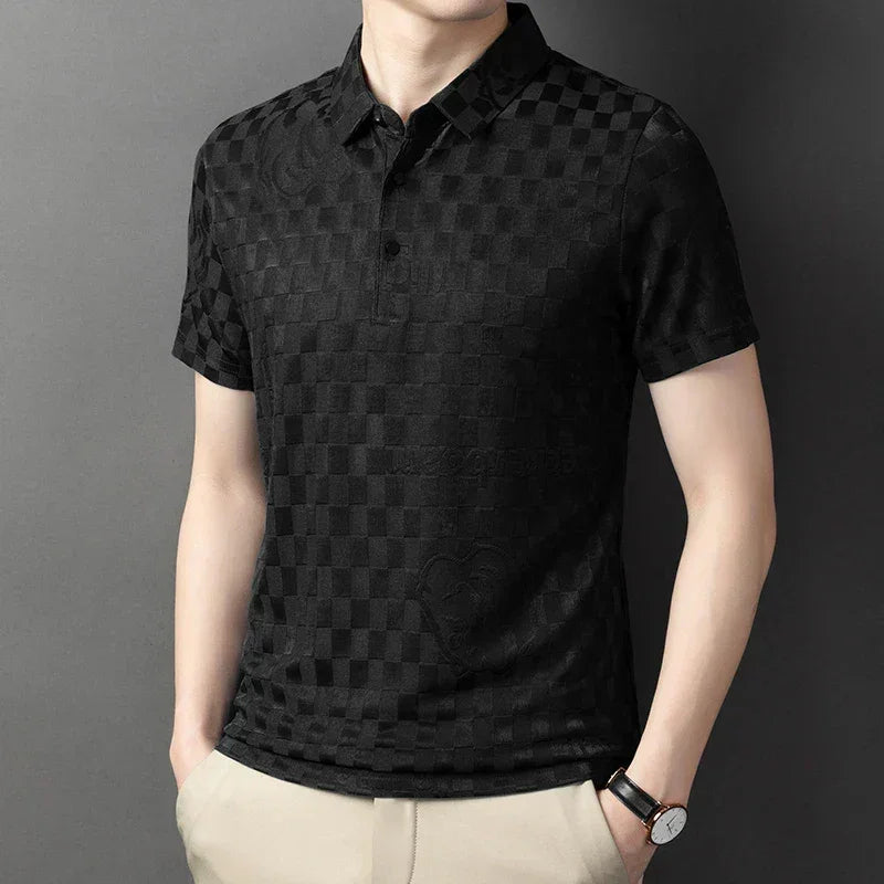 A man wearing a short-sleeved, black checkerboard pattern polo shirt with a tailored fit, beige pants, and a watch stands against a plain gray background with his hands in his pockets.