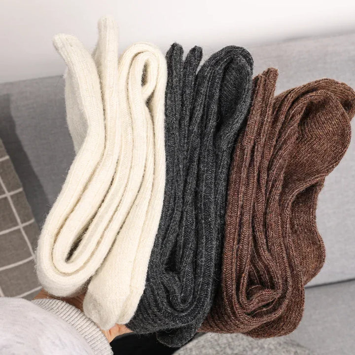 A hand holds three pairs of breathable socks made from a luxurious cashmere cotton blend in cream, dark gray, and brown, with a gray couch and checked-pattern pillow in the background.