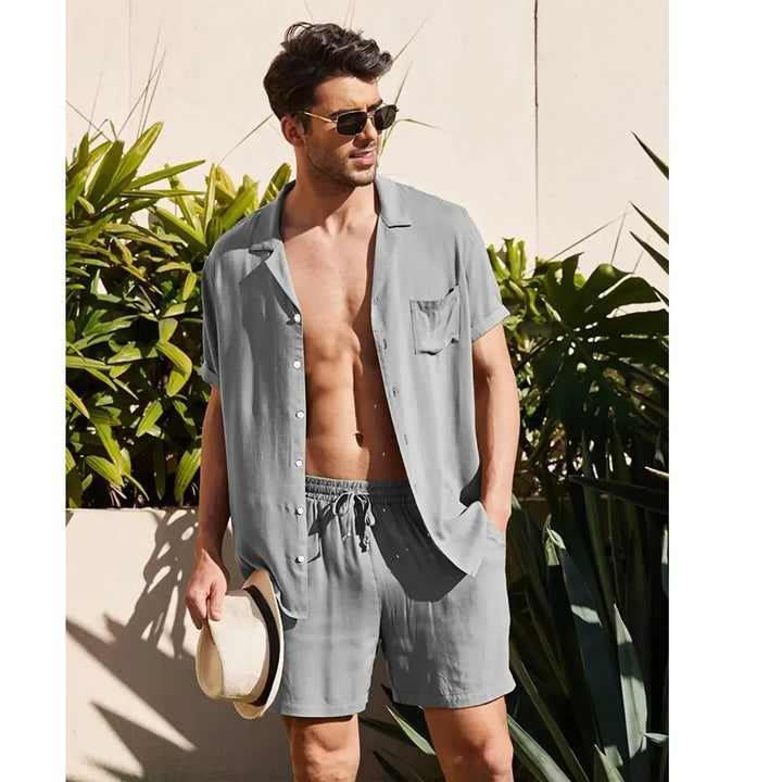 Man in light gray short sleeve shirt and shorts, sunglasses, holding hat, standing by tropical plants