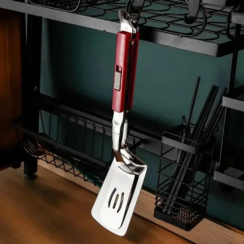 A metal spatula with a red handle, essential for BBQ season, hangs from a black kitchen rack above a wooden countertop. The spatula has slots for draining and is neatly stored with other kitchen utensils nearby.