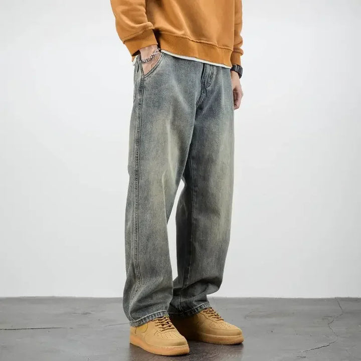 A person wearing a brown sweatshirt, classic fit stone washed jeans, and tan sneakers stands with hands in pockets on a gray floor against a plain white background.