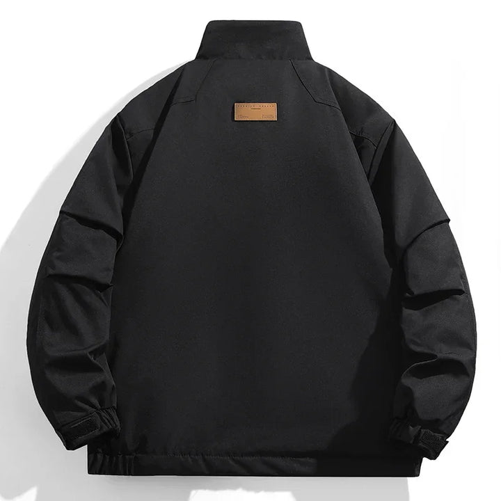 Black windproof jacket viewed from the back, featuring a high collar, adjustable cuffs, and a small rectangular brown patch near the upper center of the back. The background is white.