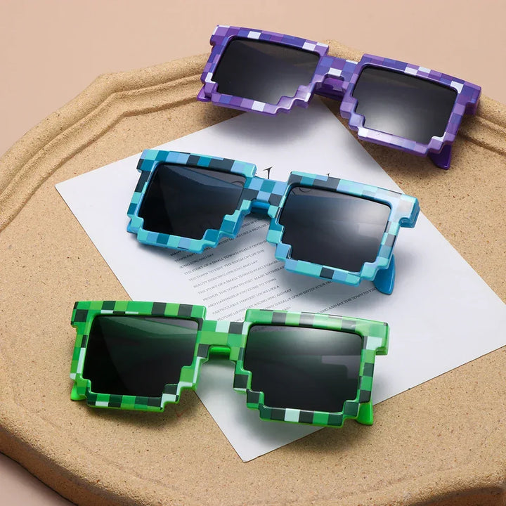 Three pairs of pixelated sunglasses in purple, blue, and green with chunky, block-like frames channel a retro-gaming aesthetic as they rest on a beige tray with white paper underneath.