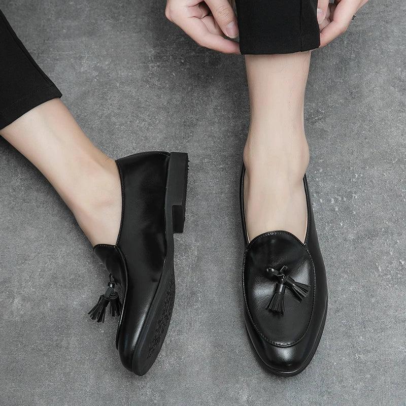 A person wearing black leather tassel loafers with black pants, sitting on a grey textured floor. One shoe, known for its cushioned insole, is on while the other foot is bare with the shoe held nearby.