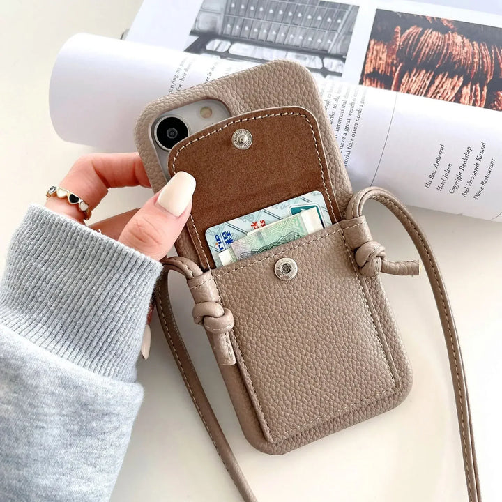 A hand holds a taupe leather phone case with a front pocket card holder containing cards. The case features a snap button closure, a detachable lanyard, and is set against an open magazine on a white surface in the background.