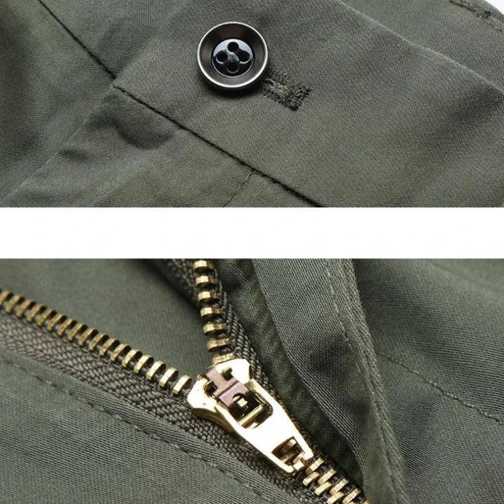Close-up view of olive green pants showing a black button at the waistband and a partially unzipped brass zipper below, highlighting the tailored fit and versatile style.