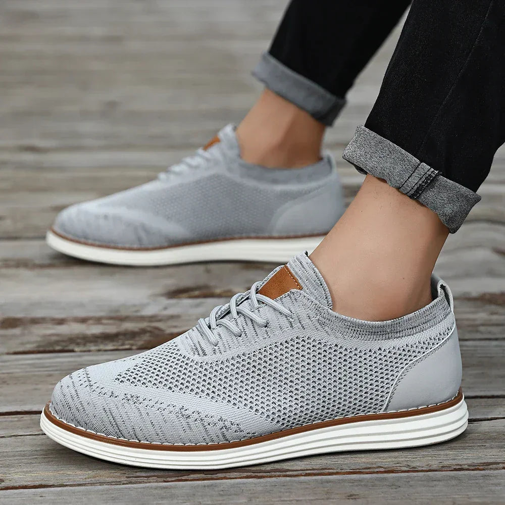 Gray lightweight sneakers with white soles are worn by someone standing on a wooden surface. The person is sporting black pants with rolled cuffs, showing off their ankles for a relaxed, casual style.