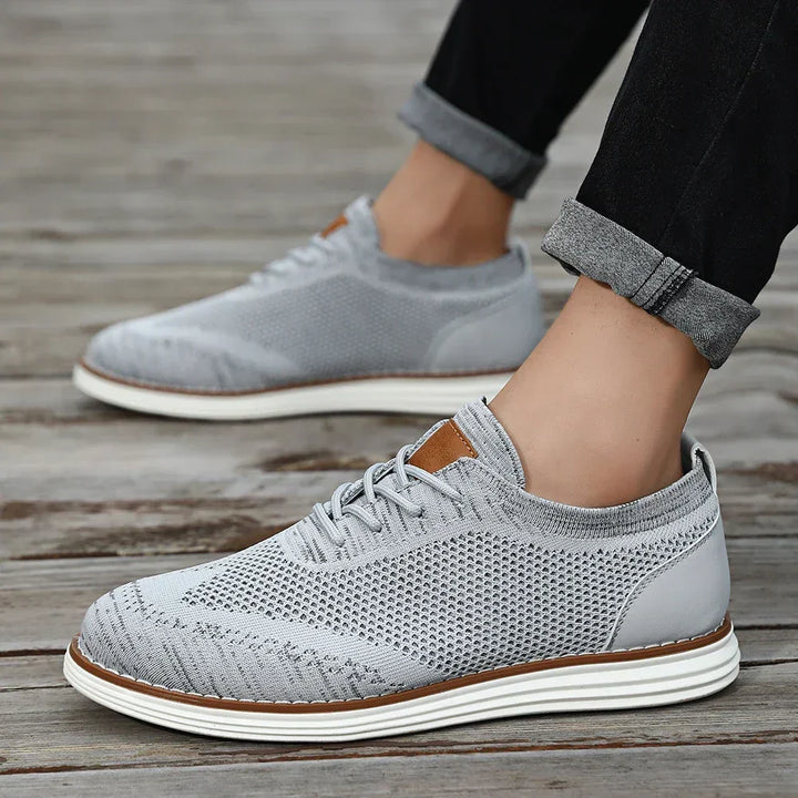 Gray lightweight sneakers with white soles are worn by someone standing on a wooden surface. The person is sporting black pants with rolled cuffs, showing off their ankles for a relaxed, casual style.