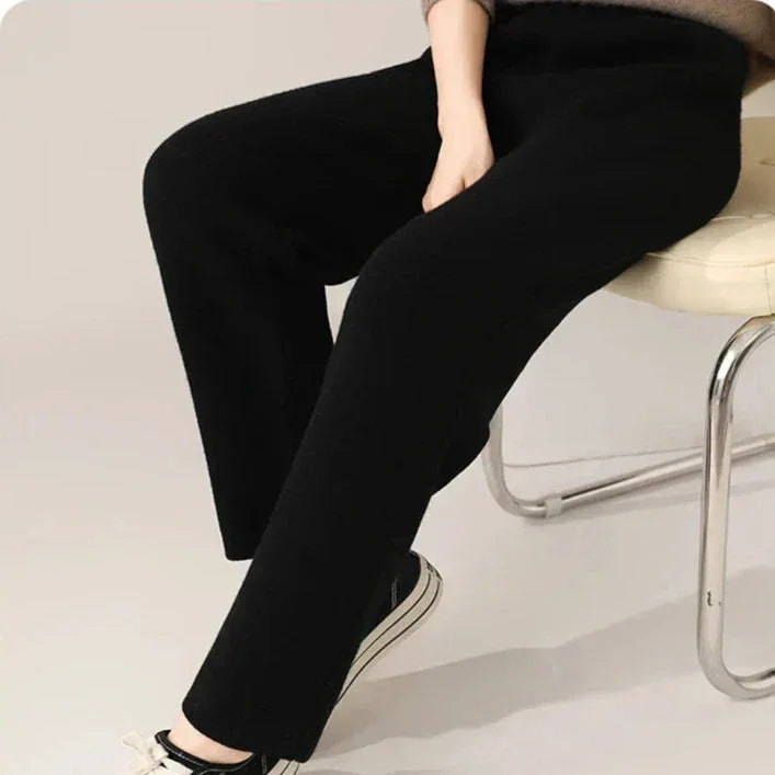 A person sits on a light-colored cushioned chair, wearing loose black pants featuring ultra soft insulation and black-and-white sneakers. Only the lower half of the body is visible.
