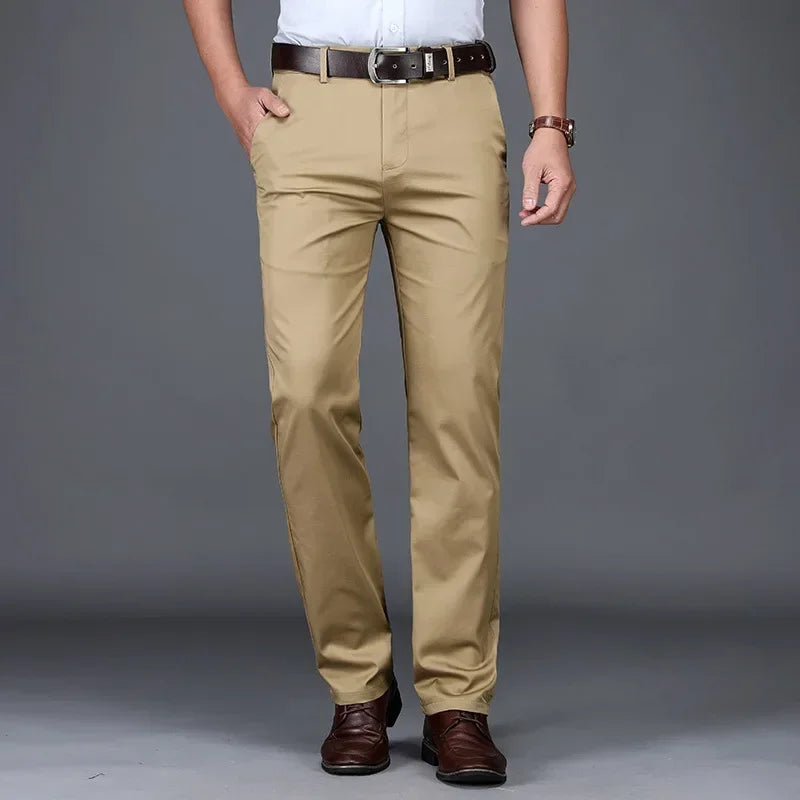A man stands against a gray background wearing tan tailored fit pants, a white shirt, a dark brown belt, brown shoes, and a watch. His hands are in his pockets. His head is not visible in the image.
