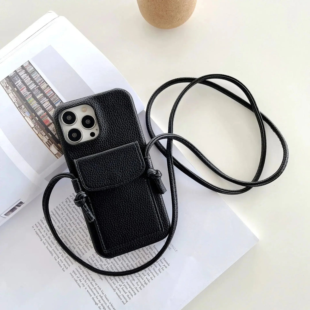 A black textured leather phone case with a card holder, small pouch, and detachable lanyard rests on an open book atop a white surface. A round, beige object is partially visible at the top of the image.
