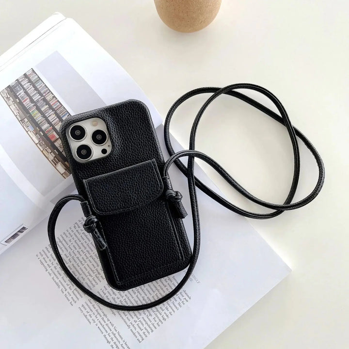 A black textured leather phone case with a card holder, small pouch, and detachable lanyard rests on an open book atop a white surface. A round, beige object is partially visible at the top of the image.