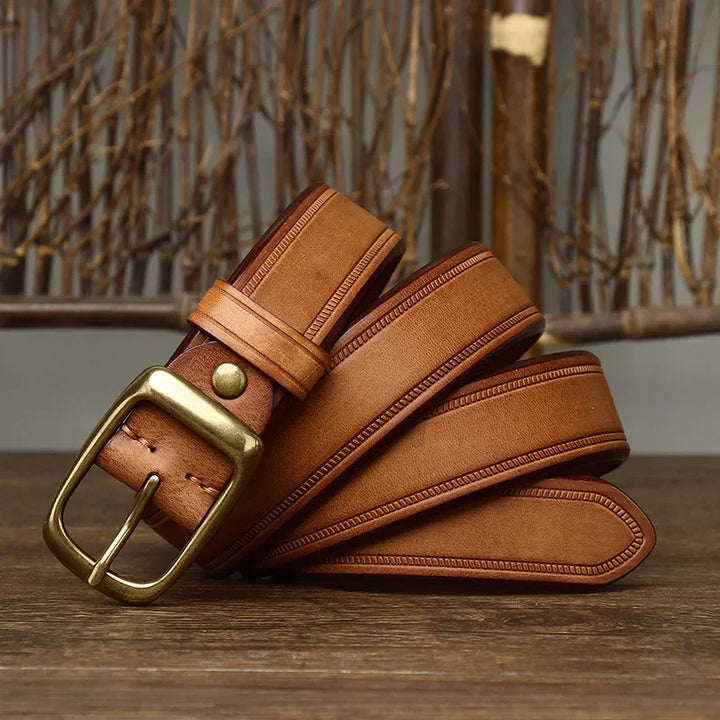 A tan cowhide belt with a brass buckle is coiled on a wooden surface, with a rustic, twig-style background visible behind it.