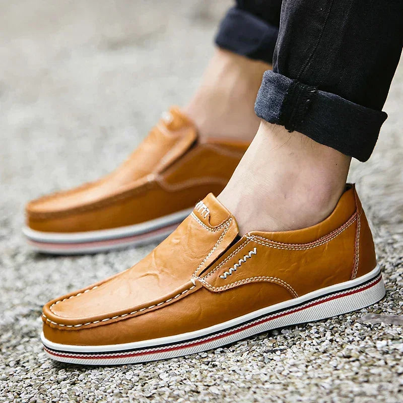 A person wearing light brown leather boat-style loafers with white and red-striped soles stands on a gravel surface. The person’s jeans are rolled up at the ankles.