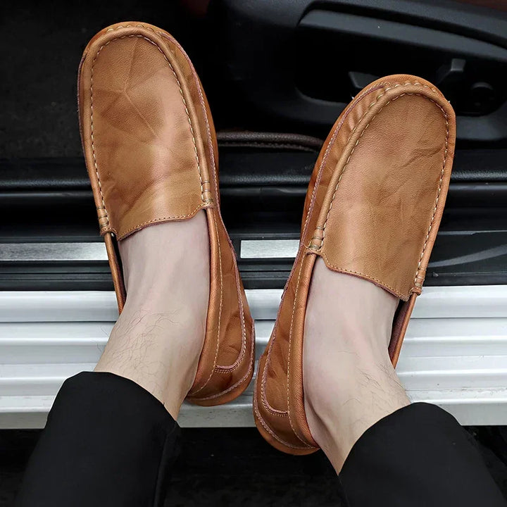 A person wearing genuine leather loafers with visible stitching and a cushioned insole is seated with feet resting on a white surface, dressed in black pants.
