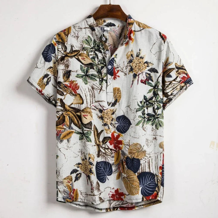 A short-sleeved button-up Vintage Shirt Men with an oversized fit, featuring a colorful tropical floral and leaf pattern in brown, yellow, green, navy, and red on a light background, hanging on a wooden hanger against a white wall.