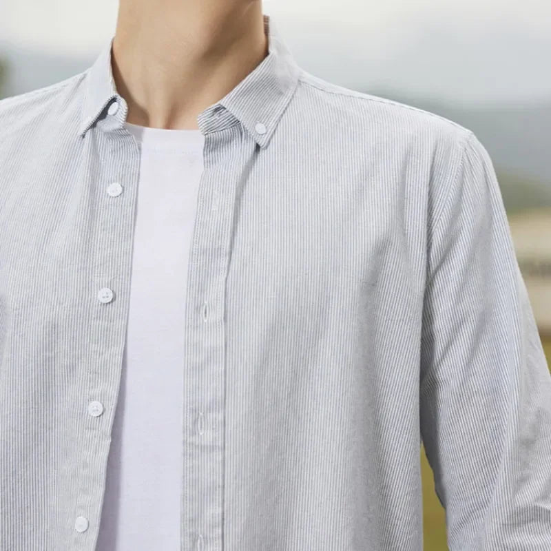 A person wearing a classic pinstripe cotton button-up shirt over a white t-shirt, photographed from the shoulders to the chest, with an outdoor background blurred.