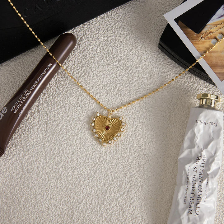 Rovelli gold heart pendant necklace with crystals, styled with hand cream and lip tint flat lay