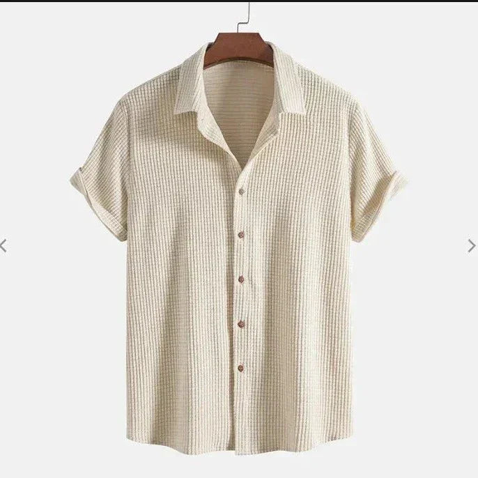 A beige, short-sleeved Bayside Shirt with a textured button-up design and collar, crafted from breathable cotton, displayed on a hanger against a plain white background.