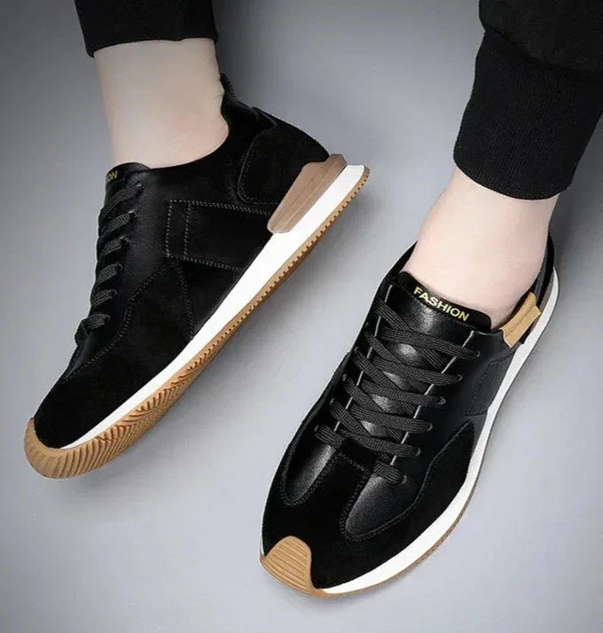 A person wearing black sneakers with white soles, brown toe caps, and the word FASHION printed on the tongue, shown from above against a light gray background. These shoes offer arch support for enhanced comfort.