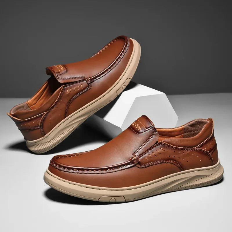 A pair of men’s cowhide leather slip-on shoes with beige soft soles, displayed against a gray background with one shoe propped up on a white geometric block.