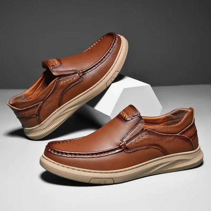 A pair of men’s cowhide leather slip-on shoes with beige soft soles, displayed against a gray background with one shoe propped up on a white geometric block.