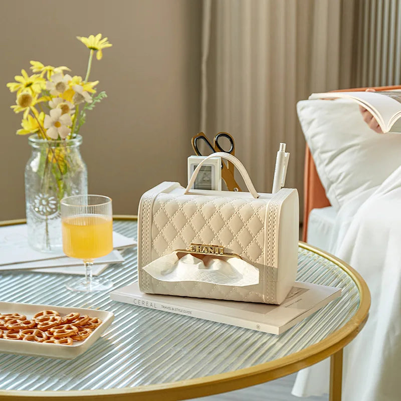 A quilted fragrance tissue box holder with a gold accent and compartments for pens and scissors sits on a coffee table beside a glass of orange juice, a vase of yellow flowers, and pretzels, adding charm to your home decor near the neatly made bed.