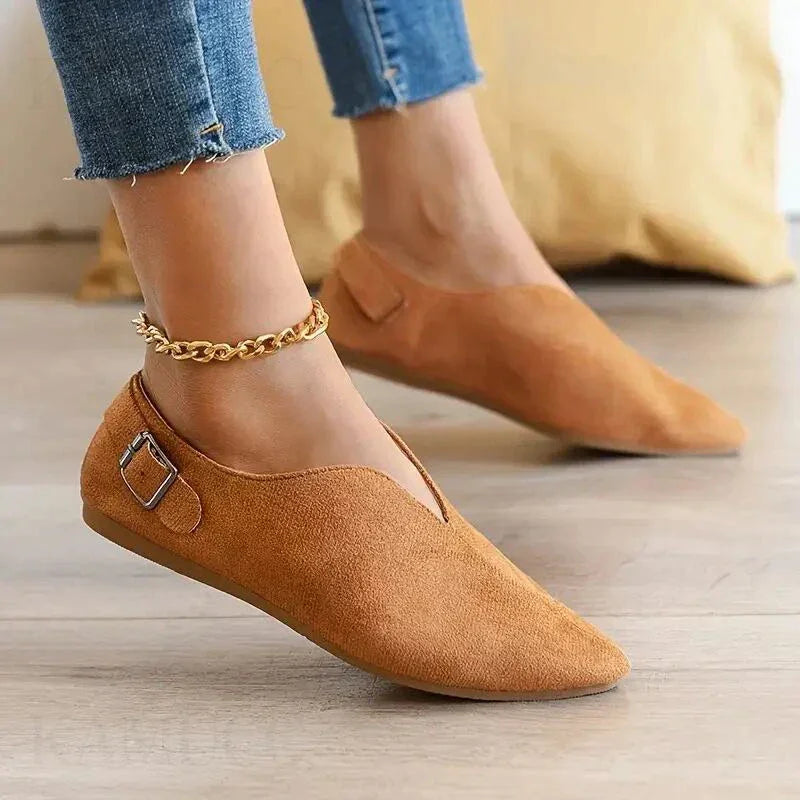 A person wearing tan suede pointed toe shoes with a cushioned insole, buckle detail, and ankle chain, paired with frayed hem blue jeans, stands on a light wooden floor.