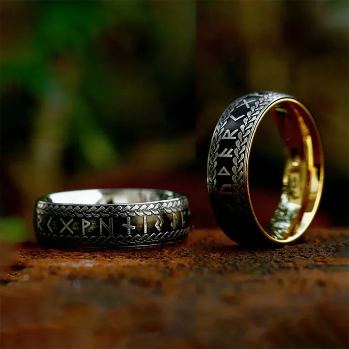Two Viking Rune Rings with silver runic engravings and intricate patterns are displayed on a rustic surface. One of these stainless steel rings lies flat while the other stands upright, revealing a gold-colored interior. The background is softly blurred.