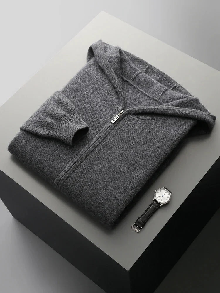 A neatly folded dark gray men wool cardigan with a hood is placed on a light gray platform. A silver wristwatch with a black band lies next to it. The scene is minimal and well-lit.