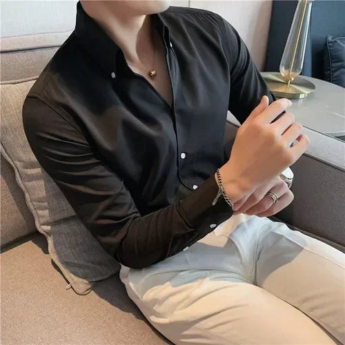 A person wearing a black wrinkle resistant shirt and white pants sits on a sofa, with arms partially crossed and hands resting on their lap. A lamp and table are visible in the background.