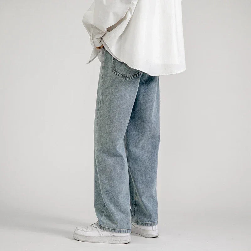 A person wearing light blue premium denim relaxed fit jeans, a white oversized shirt, and white sneakers stands against a plain light gray background, facing sideways with hands partially in pockets.