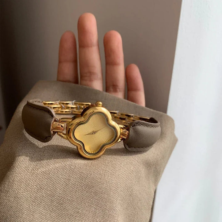 A luxury gold-plated clover dial watch with a genuine leather strap rests on a person’s raised hand, partially covered by a beige sleeve.