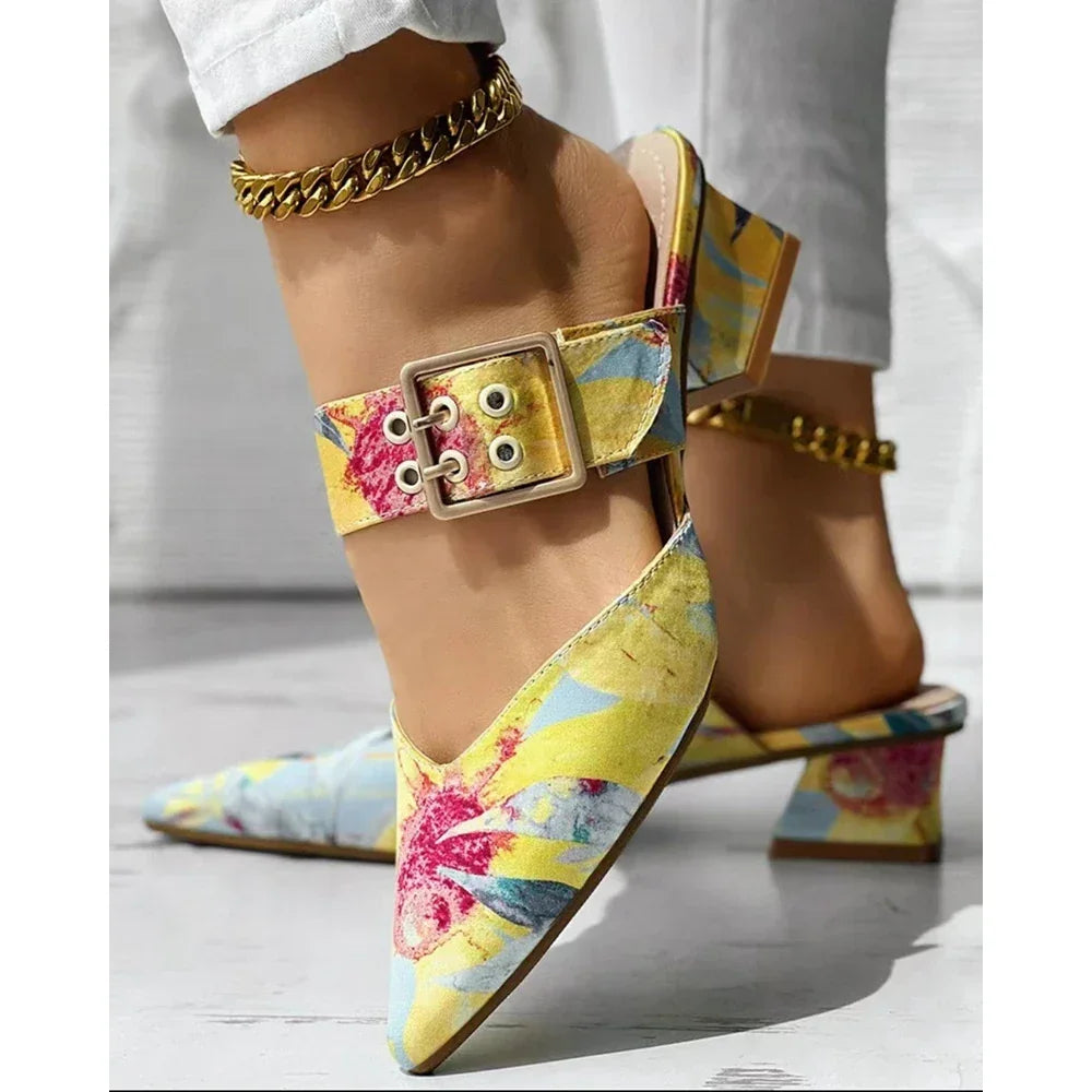 Close-up of a person wearing white pants and colorful floral pointed-toe mules slippers with chunky gold chain anklets and large buckle straps. These summer shoes feature a square heel, yellow base, and red and blue floral patterns.