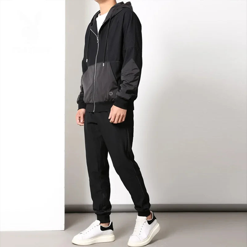 A person stands indoors wearing a black and gray zipper hoodie and jogger pants, creating a sleek activewear set paired with white sneakers. The background is plain with white and gray walls, and their head is not visible in the image.