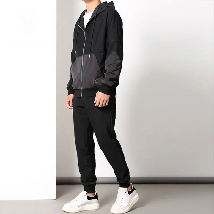 A person stands indoors wearing a black and gray zipper hoodie and jogger pants, creating a sleek activewear set paired with white sneakers. The background is plain with white and gray walls, and their head is not visible in the image.
