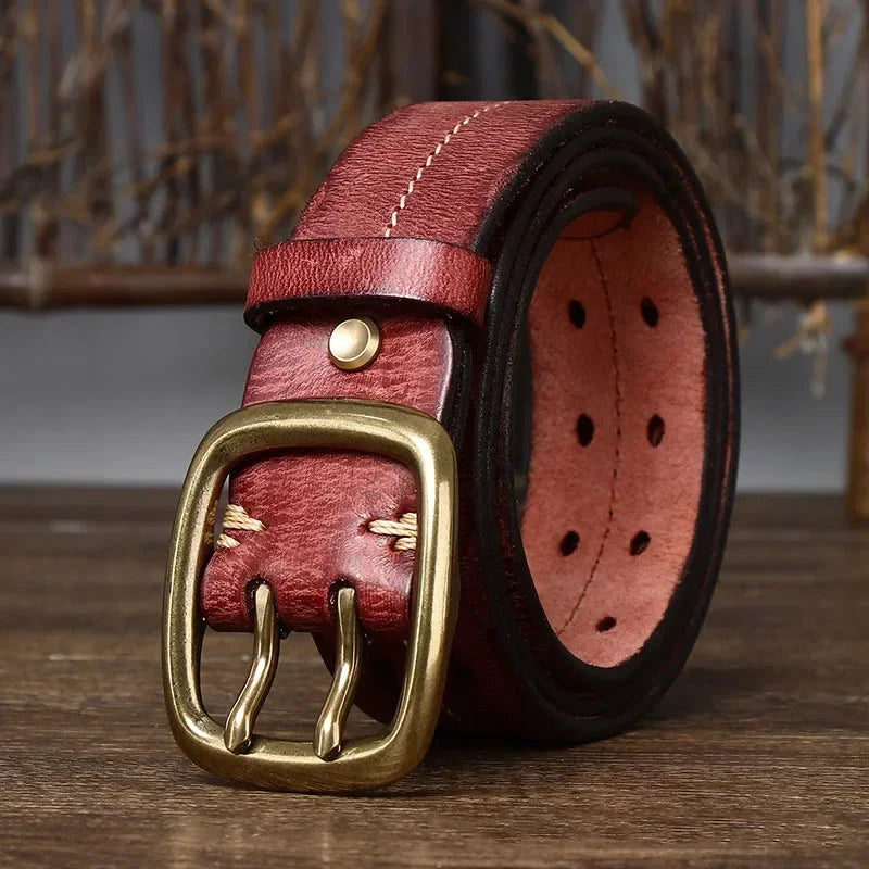 A thick, reddish-brown genuine leather belt with beige stitching and a large, square, gold-colored double prong buckle is rolled up and displayed on a wooden surface, showcasing its heritage craftsmanship.