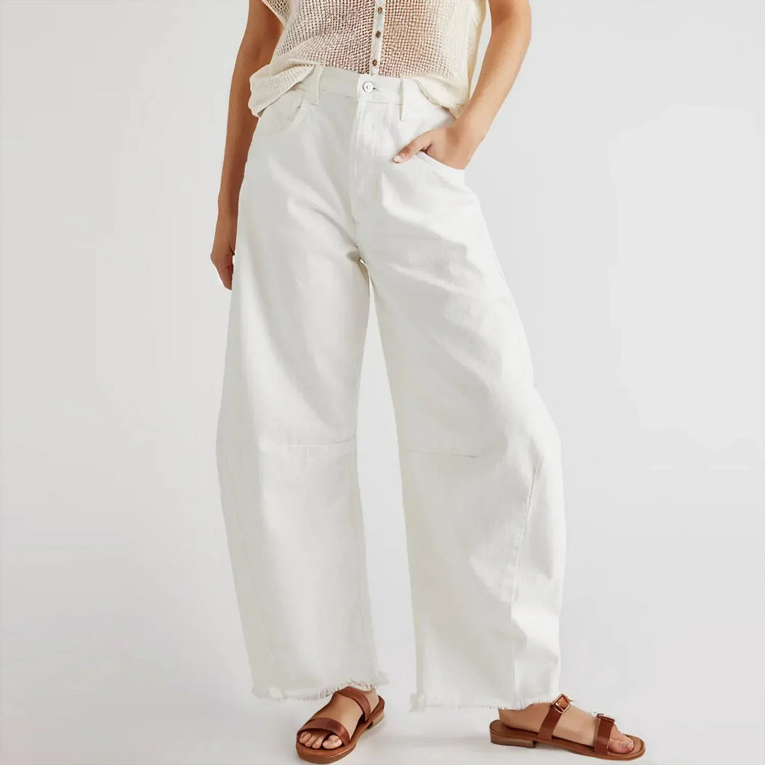 A person wearing relaxed fit white jeans, a sleeveless netted top, and brown sandals stands with one hand in their pocket against a plain white background.