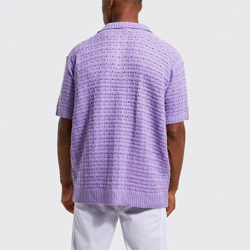A person stands facing away from the camera, wearing a light purple, short-sleeved summer shirt with a breathable knit design and white pants against a plain light background.
