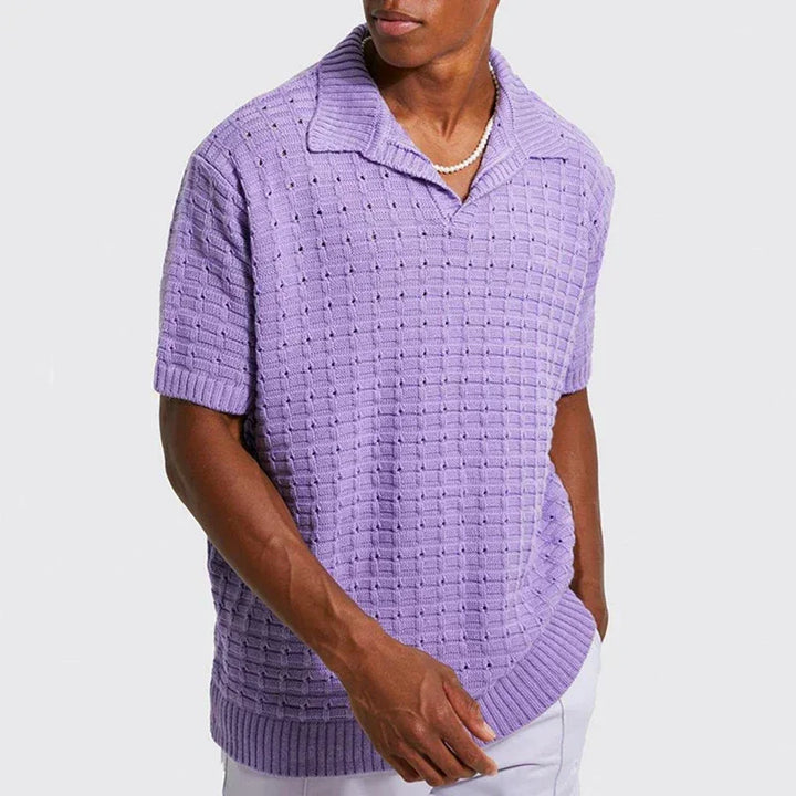 A man wearing a breathable knit design, short-sleeve light purple summer shirt with a collar and white pants, accessorized with a pearl necklace, stands with one hand in his pocket against a light gray background.