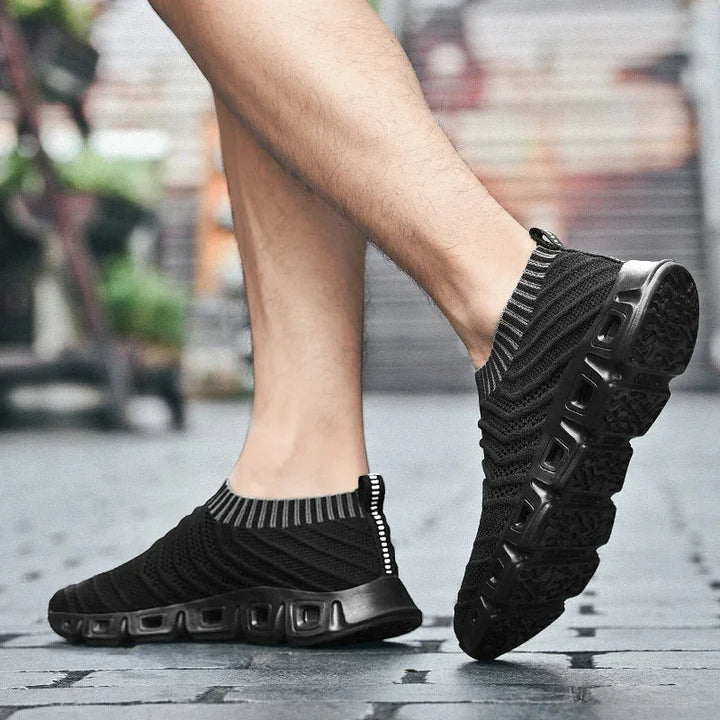 A person wearing black slip-on athletic shoes with a sock-like fit and textured soles walks on a paved surface. The knit upper features striped detail near the ankle. The background is slightly blurred with greenery and a building.