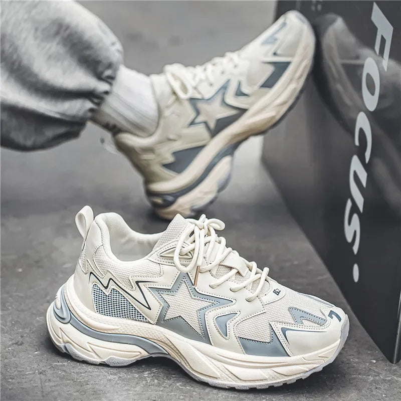 A person wearing white and light gray chunky sneakers with star patterns and a durable rubber outsole sits on the ground, one foot propped against a black box labeled Focus. They are dressed in light gray sweatpants.