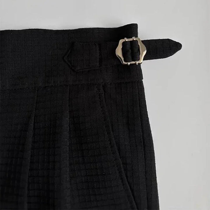 Close-up of the waistband of black luxury tailored pants with a silver buckle and strap detail on the side, set against a light background.