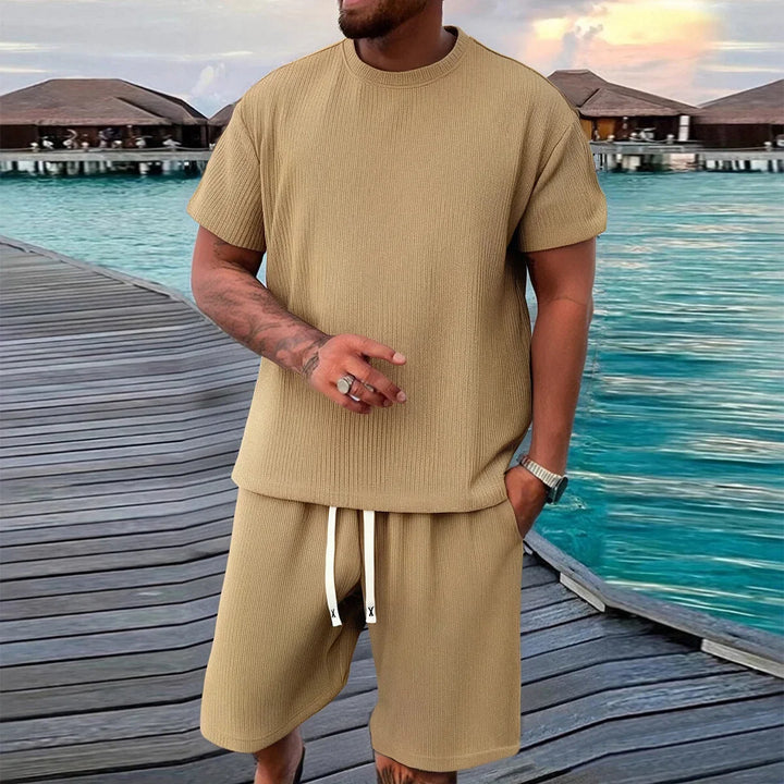 Man in beige lounge set standing on wooden dock by blue water, Rovelli fashion style