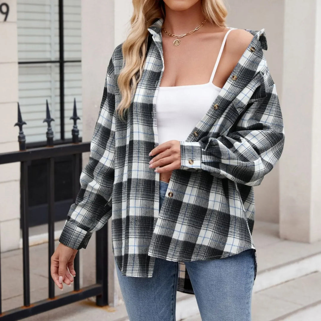 A woman stands outdoors wearing a black and white plaid button-down shirt in an oversized fit over a white tank top and blue jeans. She has wavy hair and a gold necklace. Her face is not visible.