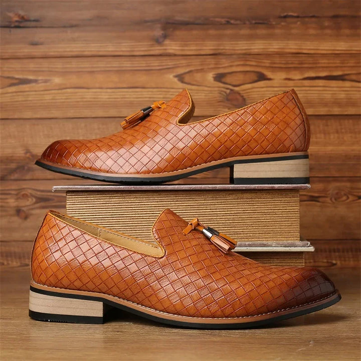 A pair of brown real leather loafers with tassels, featuring a basketweave pattern, is displayed against a wooden background. One shoe sits atop two stacked books, while the other rests on the surface below.