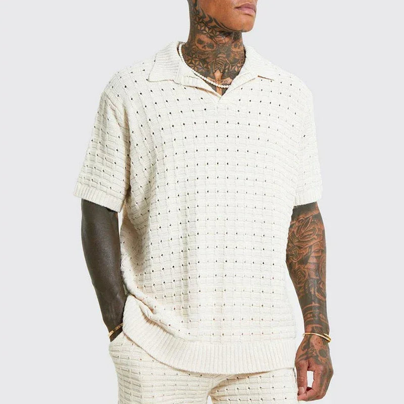 A person with tattoos on their neck and arms wears a cream-colored, short-sleeve summer shirt with a collar and matching knit pants. The shirt features a breathable knit design with a textured, perforated pattern. The background is plain light gray.