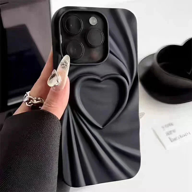 A hand holds a black phone case made of soft matte silicone with a 3D heart design in the center and a silky, draped texture. The person’s nails are adorned with floral art, adding a touch of elegance to the scene.