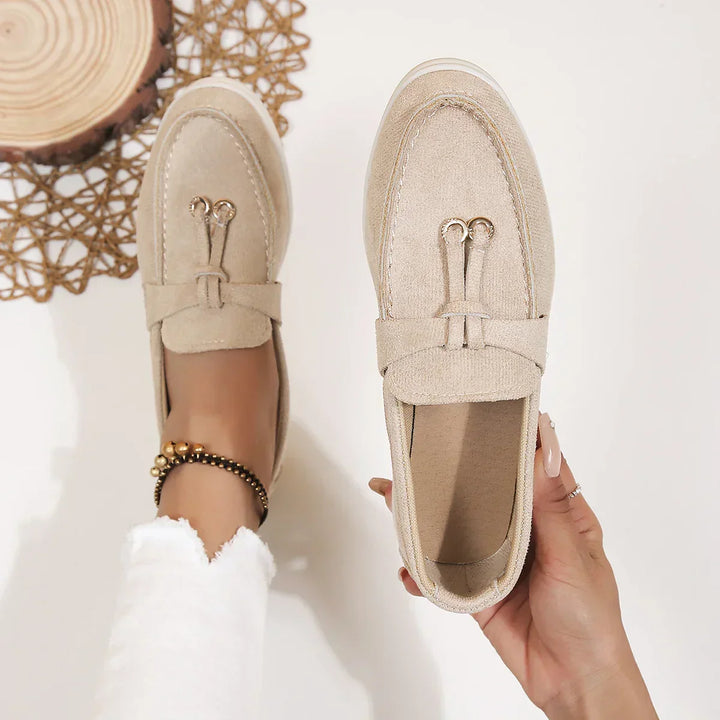 A person in white pants and a beaded anklet holds a chic moccasin while wearing its match; a woven mat and a wooden decor piece are visible in the background.