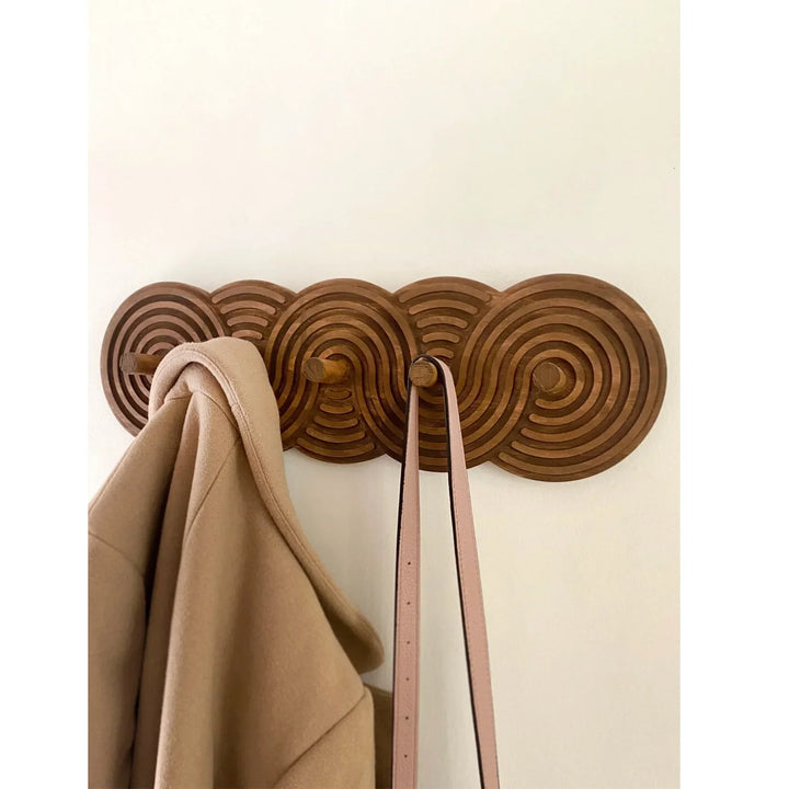 Wooden wall coat rack with geometric pattern holding a beige coat and pink belt, Rovelli